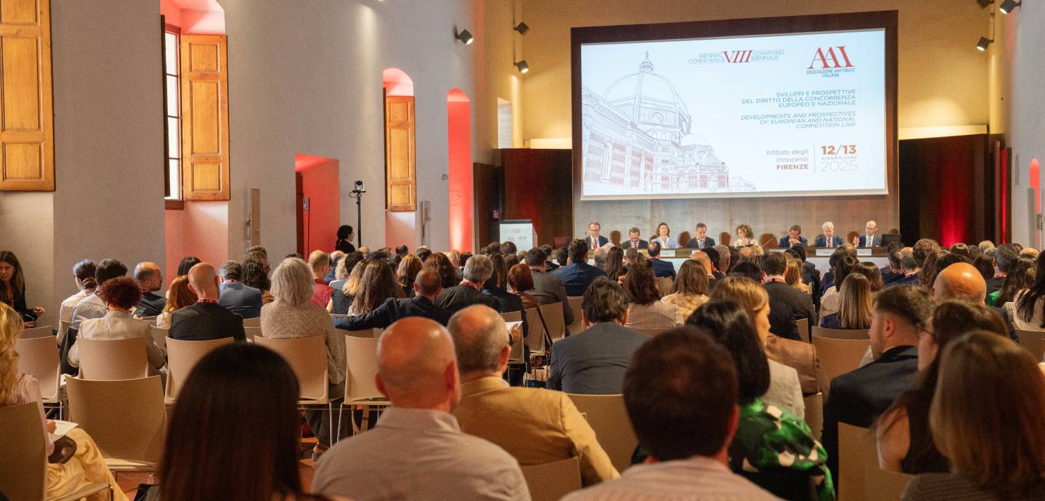 Una vasta sala conferenze, caratterizzata da un'elegante architettura storica con soffitto a volta, finestre ad arco e pareti chiare, è riempita da un pubblico numeroso seduto su sedie beige. Sul fondo della sala, un ampio palco ospita un panel di relatori seduti a un tavolo, davanti a un grande schermo. Lo schermo proietta un'illustrazione di una città storica e il logo 'BIENNIAL CONFERENCE VIII CONVEGNO BIENNALE' dell'Associazione Antitrust Italiana, con il titolo 'SVILUPPI E PROSPETTIVE DEL DIRITTO DELLA CONCORRENZA EUROPEO E NAZIONALE' e la data '12/13 GIUGNO/JUNE 2025 FIRENZE'. Le luci calde sul soffitto e gli accenti rossi creano un'atmosfera solenne e professionale.