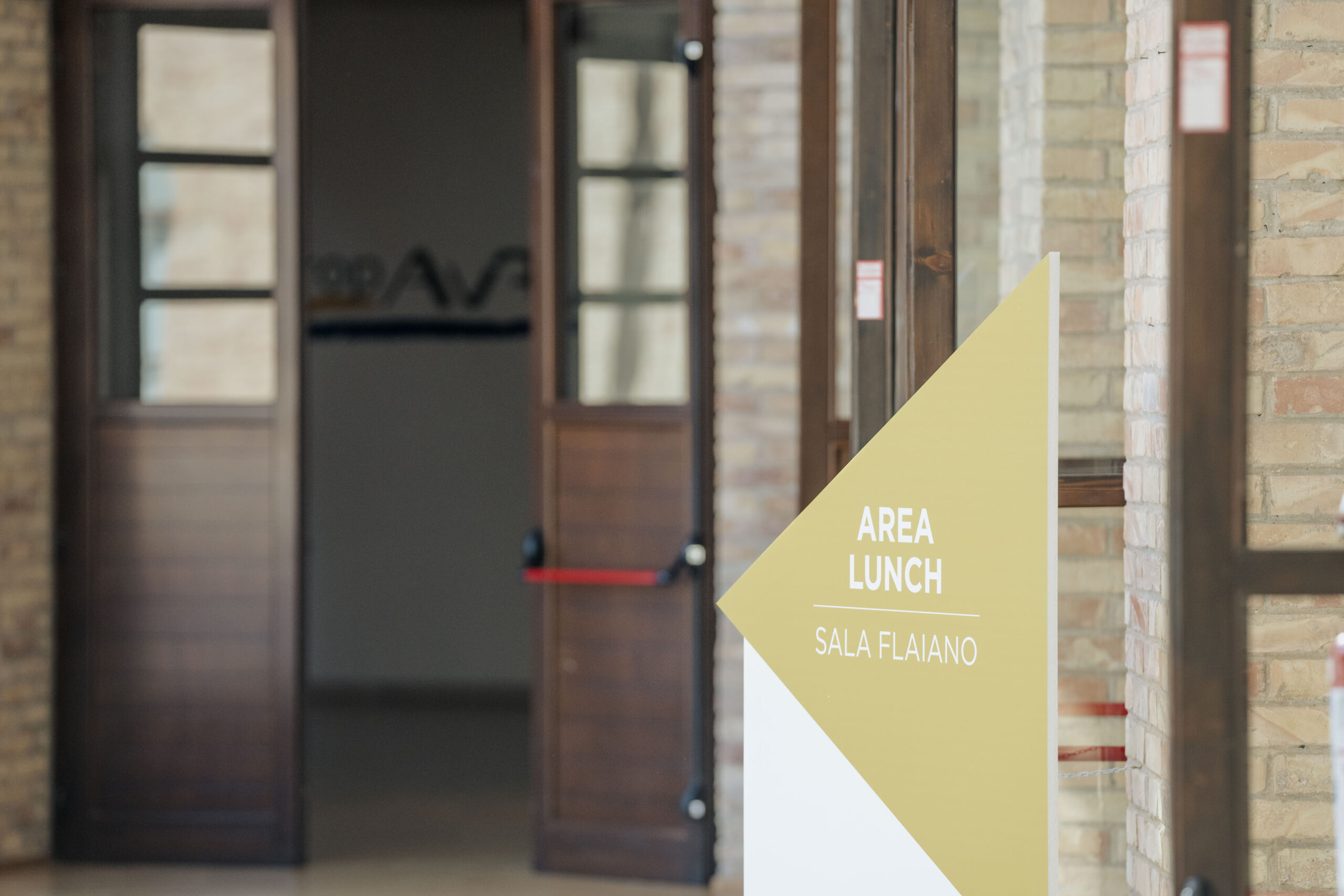 Un cartello giallo e bianco con la scritta "AREA LUNCH" e "SALA FLAIANO" indica la direzione, con delle porte in legno aperte sullo sfondo. Questo suggerisce un'area dedicata al pranzo durante un evento o una conferenza.