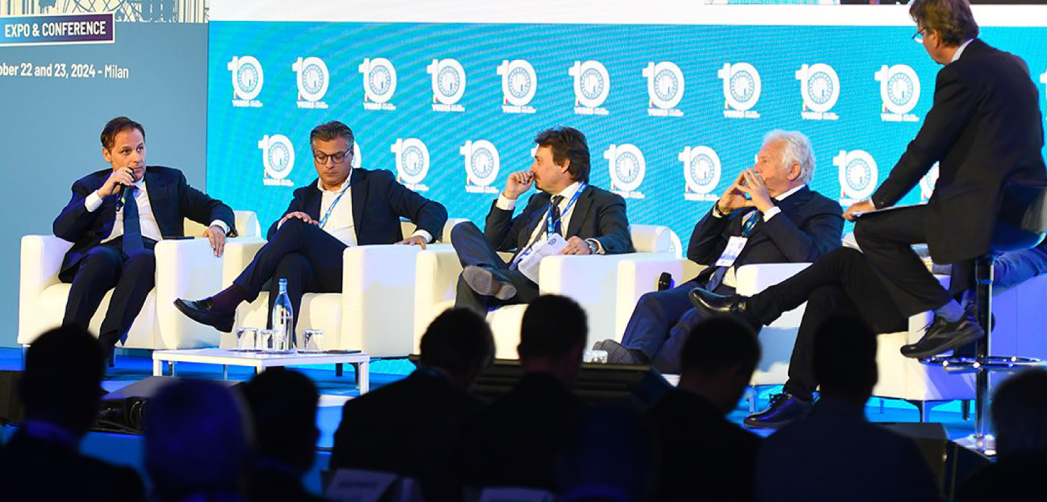 Un panel di sei uomini è seduto sul palco, accomodati su poltrone bianche e tavolini, mentre interagiscono o ascoltano, con un microfono davanti ad alcuni di loro. Dietro di loro, un grande schermo bianco mostra il logo dell'evento 'ITALIAN CRITICAL PROCESS EQUIPMENT DAYS - 10 YEARS OF AIPE' e altri loghi. In primo piano, le teste e le spalle del pubblico sono visibili, seduti nelle file dell'auditorium, suggerendo una partecipazione attenta. L'illuminazione del palco è chiara, contrastando con l'ambiente circostante meno illuminato.
