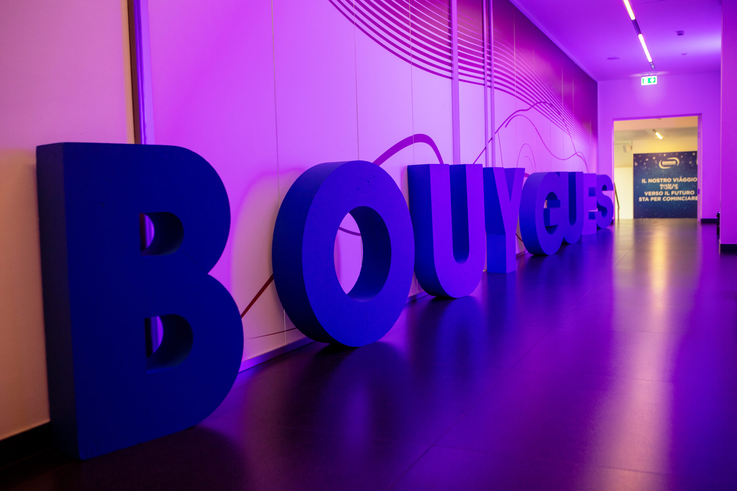Una prospettiva ravvicinata delle grandi lettere blu tridimensionali che formano la parola "BOUYGUES", posizionate su un pavimento scuro e illuminate da una luce viola-rosa. Sullo sfondo, un corridoio con una parete chiara decorata con linee ondulate e una scritta luminosa in fondo: "IL NOSTRO VIAGGIO VERSO IL FUTURO STA PER COMINCIARE".