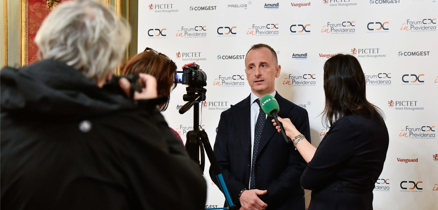 Un uomo in completo scuro e cravatta, con un microfono verde con logo 'AKSA' vicino alla bocca, sta rilasciando un'intervista. È in piedi davanti a un backdrop ripetuto con i loghi di 'PICTET Asset Management', 'COMGEST', 'Amundi', 'Vanguard' e 'Forum CDC in Previdenza'. A sinistra, si intravede la schiena di un operatore che filma con una videocamera su treppiede. A destra, un'intervistatrice tiene il microfono. L'ambiente è un'area di press conference o backstage.