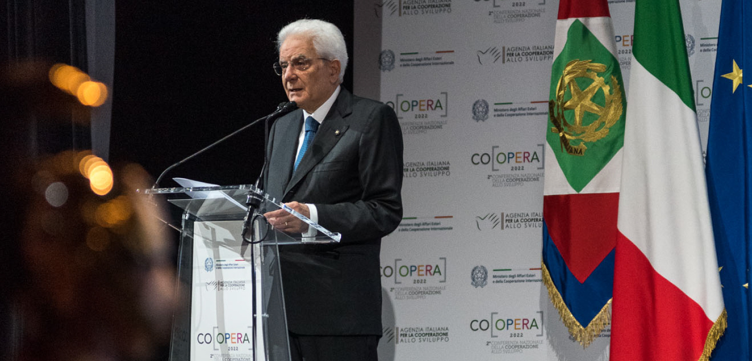 Il Presidente della Repubblica Italiana, Sergio Mattarella, è ripreso a mezzo busto mentre parla con la mano sul podio trasparente. Indossa un abito scuro e una cravatta blu chiaro. Dietro di lui, la bandiera italiana e quella dell'Unione Europea sono ben visibili, affiancate da un pannello bianco con il logo 'COOPERA 2022' e le diciture dell''Agenzia Italiana per la Cooperazione allo Sviluppo'. La sua espressione è concentrata e autorevole, tipica di un discorso ufficiale.