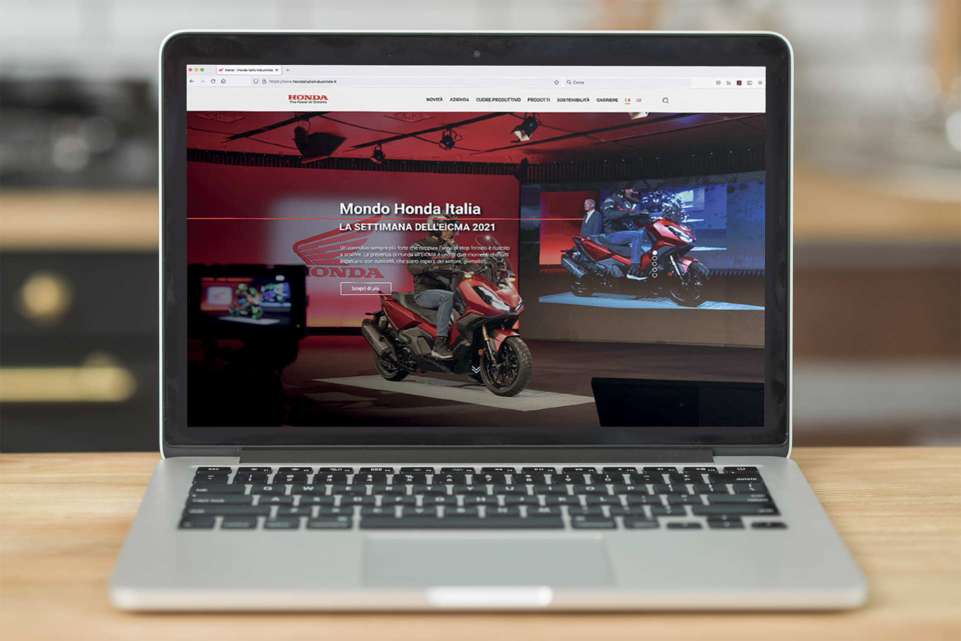 Un laptop aperto posizionato su un tavolo di legno. Lo schermo visualizza il sito web "Mondo Honda Italia", con un'immagine prominente di una motocicletta rossa e il testo "LA SETTIMANA DELL'EICMA 2021". Lo sfondo è leggermente sfocato, mostrando un ambiente interno.
