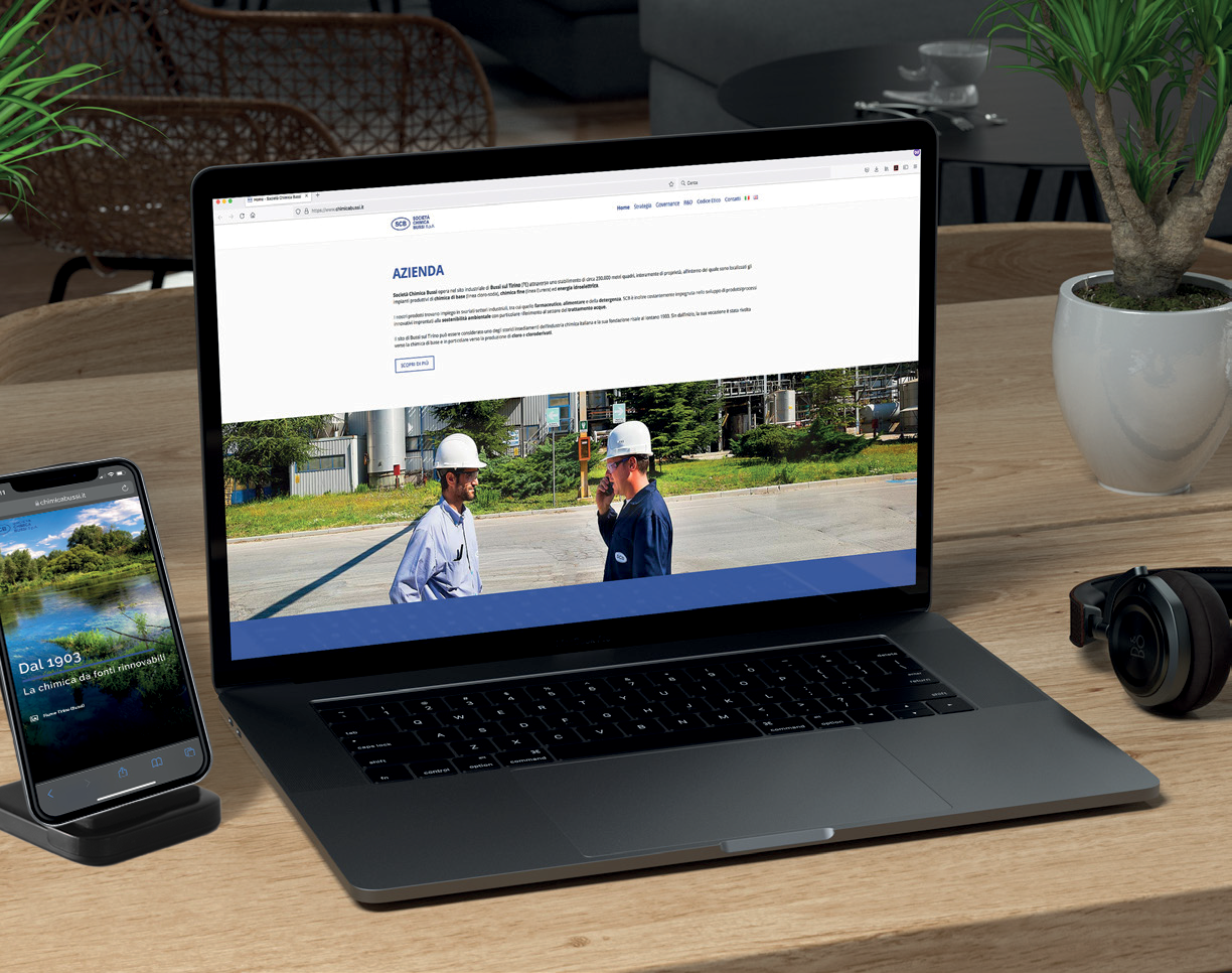 Un laptop e uno smartphone appoggiati su una scrivania di legno. Lo schermo del laptop mostra il sito web "SOGIS" con la pagina "Azienda", che include un'immagine di due lavoratori con elmetto in un impianto industriale. Lo smartphone mostra una pagina web con un paesaggio naturale e il testo "Dal 1993 La chimica da fonti rinnovabili". Cuffie e una pianta in vaso sono visibili sul lato destro.