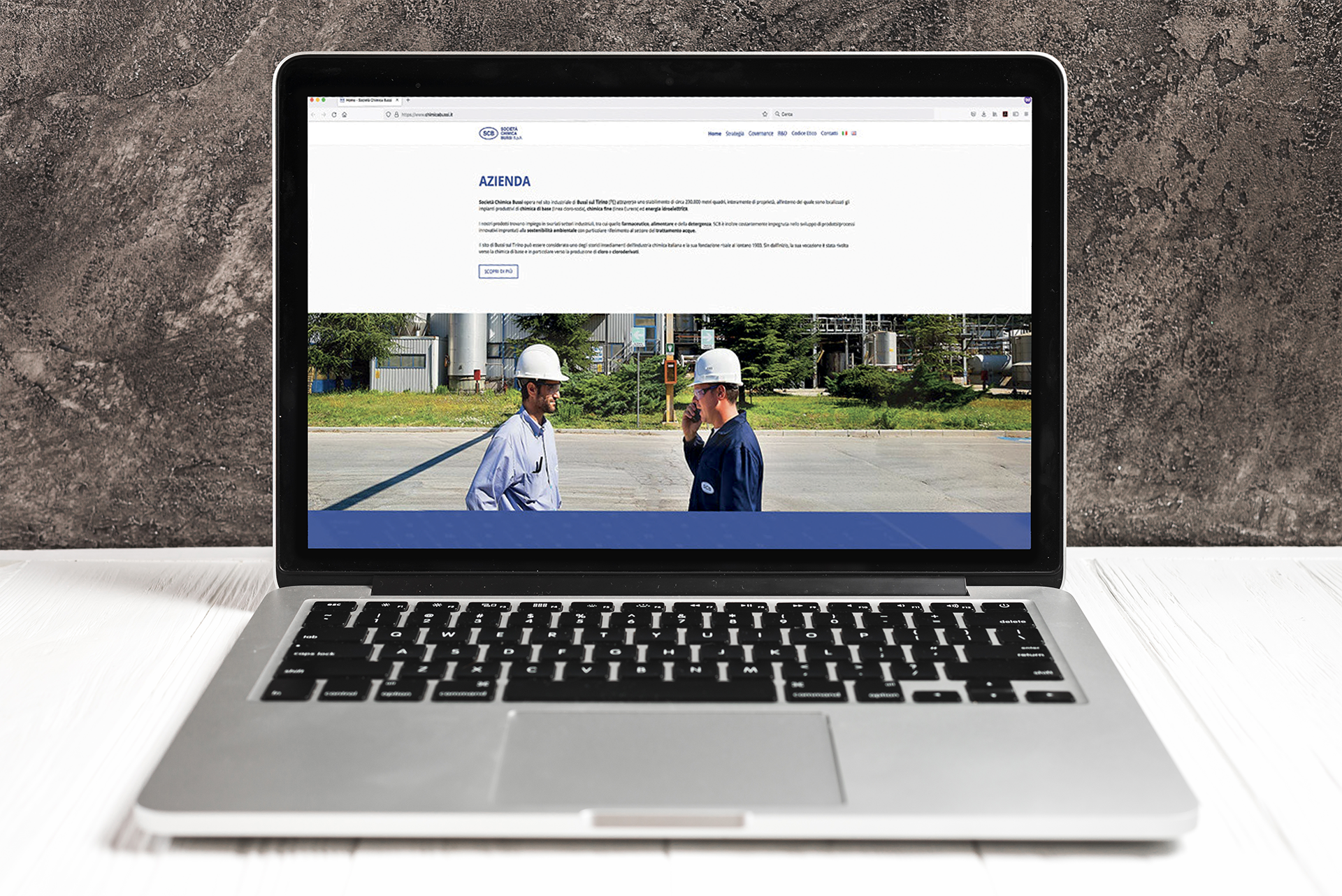 Un laptop aperto posizionato su un tavolo bianco. Lo schermo visualizza il sito web "SOGIS" sulla pagina "Azienda", con un'immagine di due uomini in elmetto che parlano in un ambiente industriale. Lo sfondo dietro il laptop è una parete grigia leggermente ruvida.