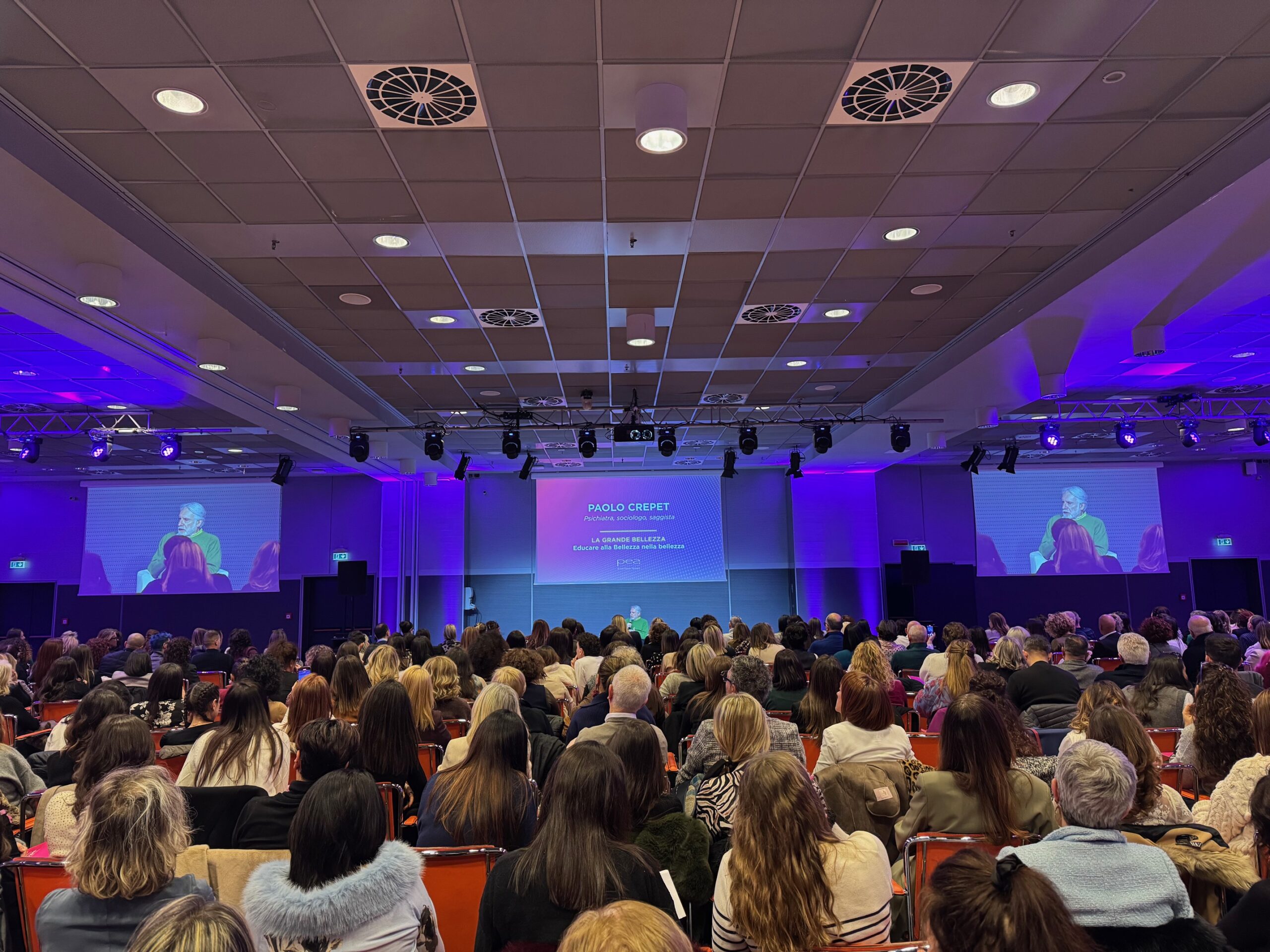 Una vista ampia di una grande sala conferenze o auditorium piena di un pubblico seduto. La sala è illuminata con luci viola d'atmosfera. Sul palco, due grandi schermi mostrano un relatore, Paolo Crepet, che è anche visibile al centro su uno schermo principale.