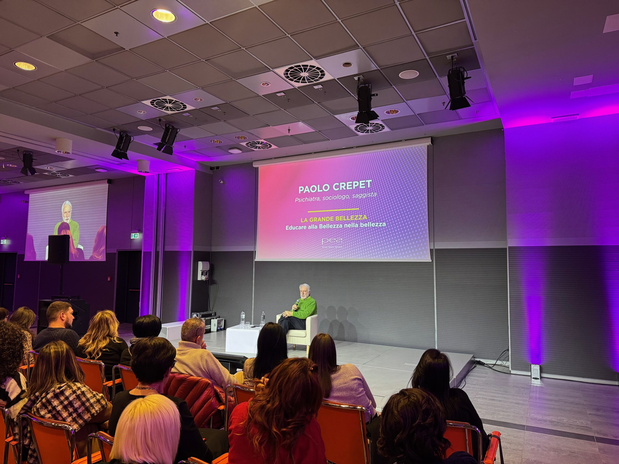 Un primo piano del palco di una conferenza. Un uomo, Paolo Crepet, seduto su una sedia, parla al pubblico. Il grande schermo dietro di lui lo identifica come psichiatra, sociologo, saggista e mostra il titolo del suo intervento: "LA GRANDE BELLEZZA Educare alla Bellezza nella Bellezza". Il palco è illuminato con luci viola.
